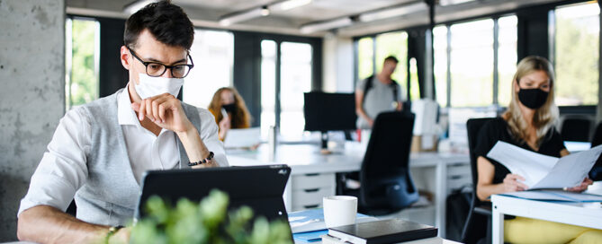 Man working on laptop wearing a mask with other colleagues.