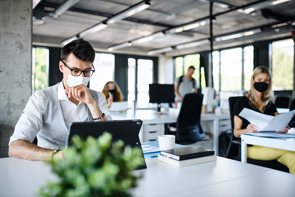 Man working on laptop wearing a mask with other colleagues.